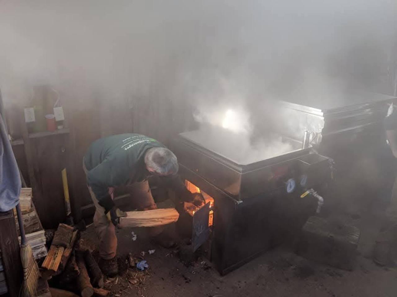 A sugar maker loading split firewood into the glowing firebox beneath the steaming evaporator at Knolltop Farm