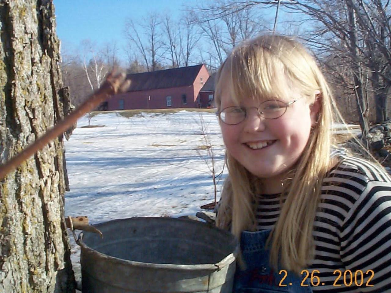 A smiling young helper beside a sap bucket on a maple tree, in a photo dated February 26, 2002
