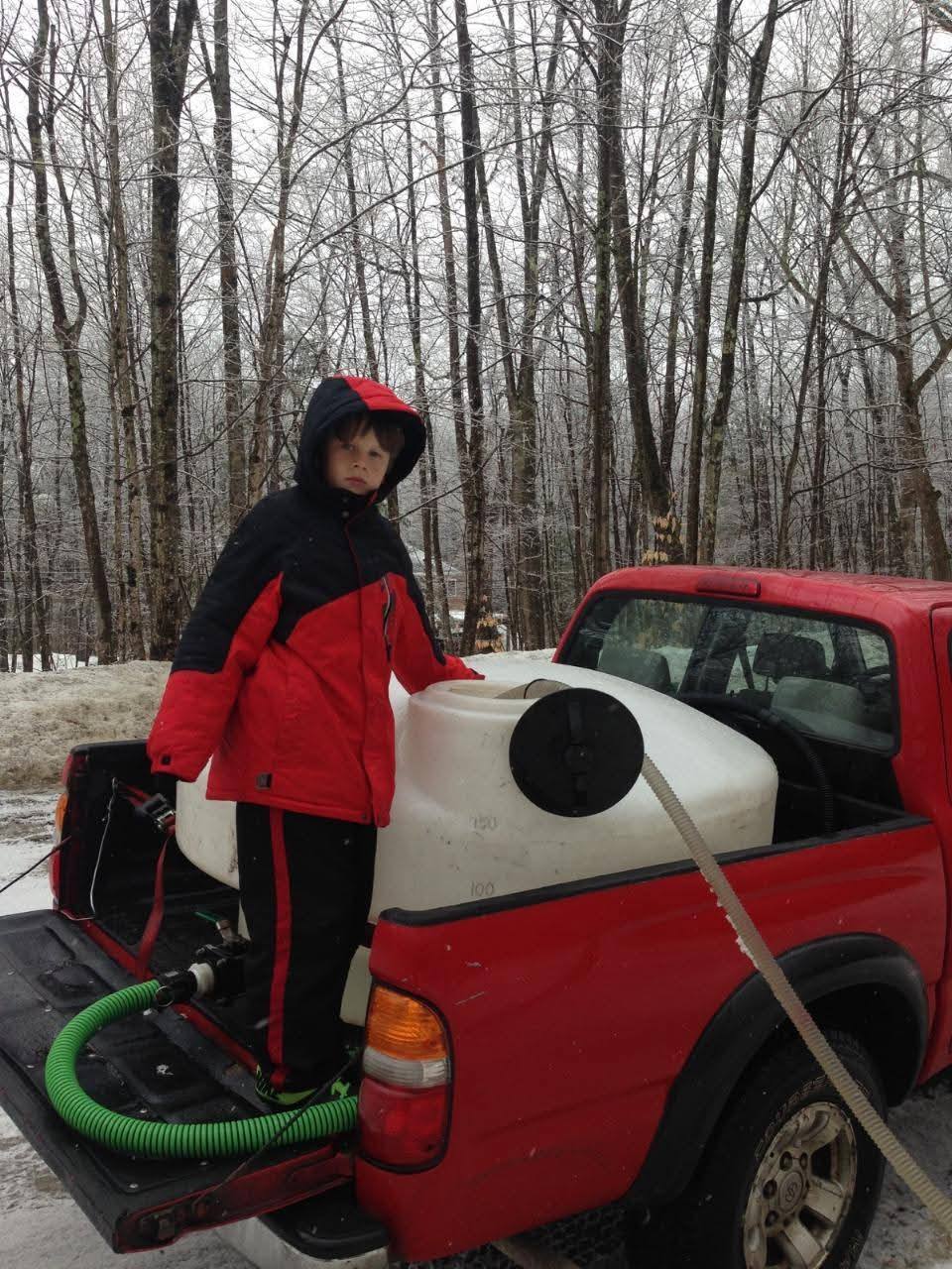 A young helper beside a sap collection tank in the bed of a red pickup in the snowy woods